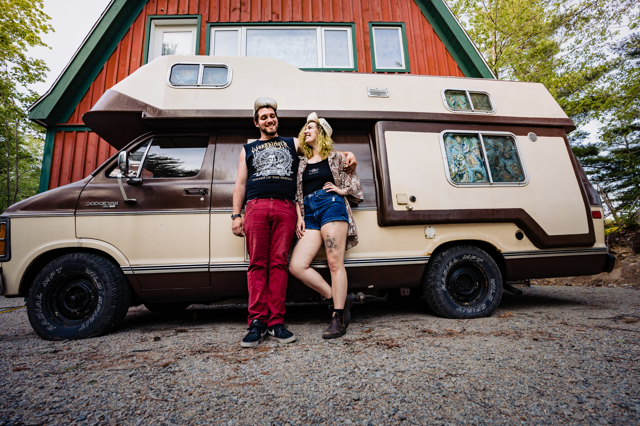 A couple stand together laughing with arms around one another in front of a hippe brown and beige van