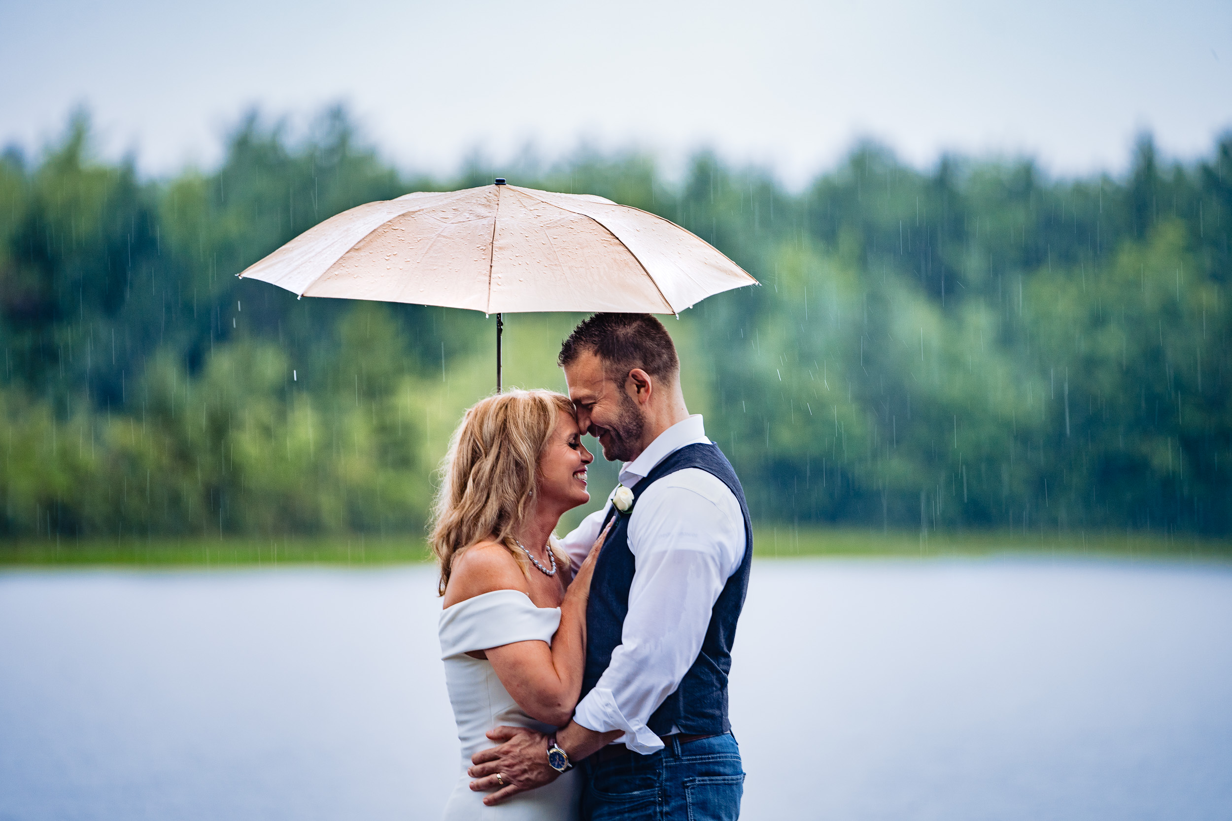 A bride and groom stand together on a dock in the rain laughing with an umbrella over them