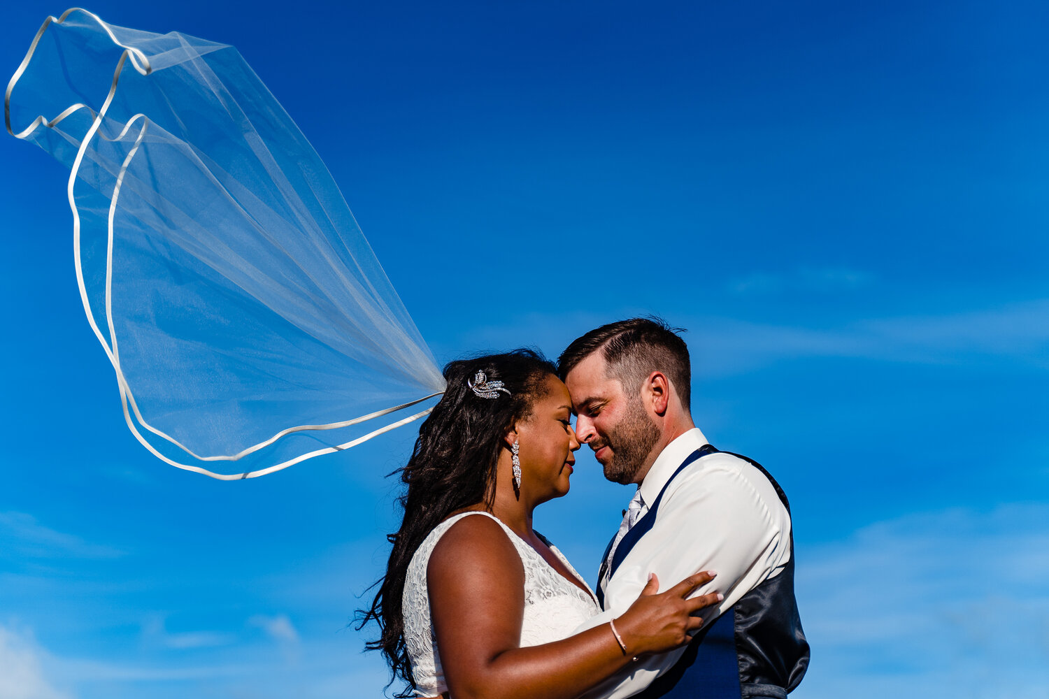 Bride and groom stand facing one another with blue sky behind them and the bride's veil blowing in the wind behind her