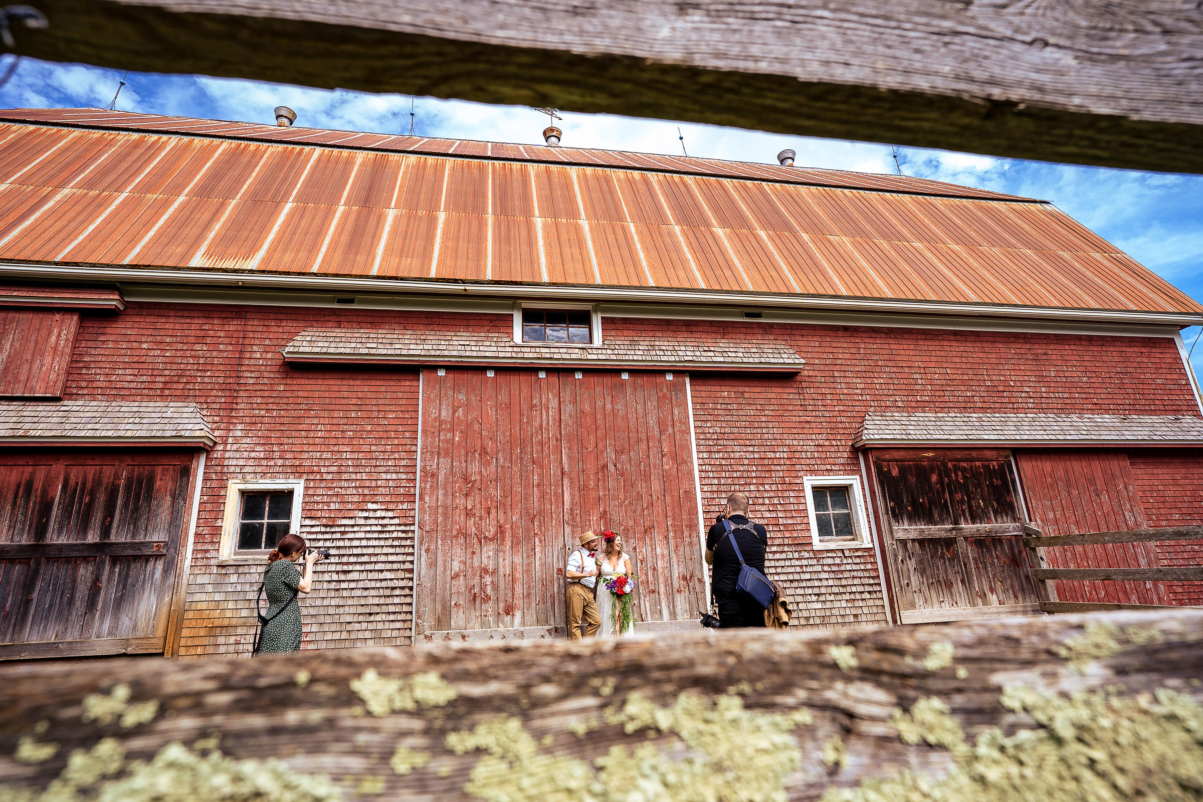 Two photographers photograph a bride and groom in front of a rustic red barn