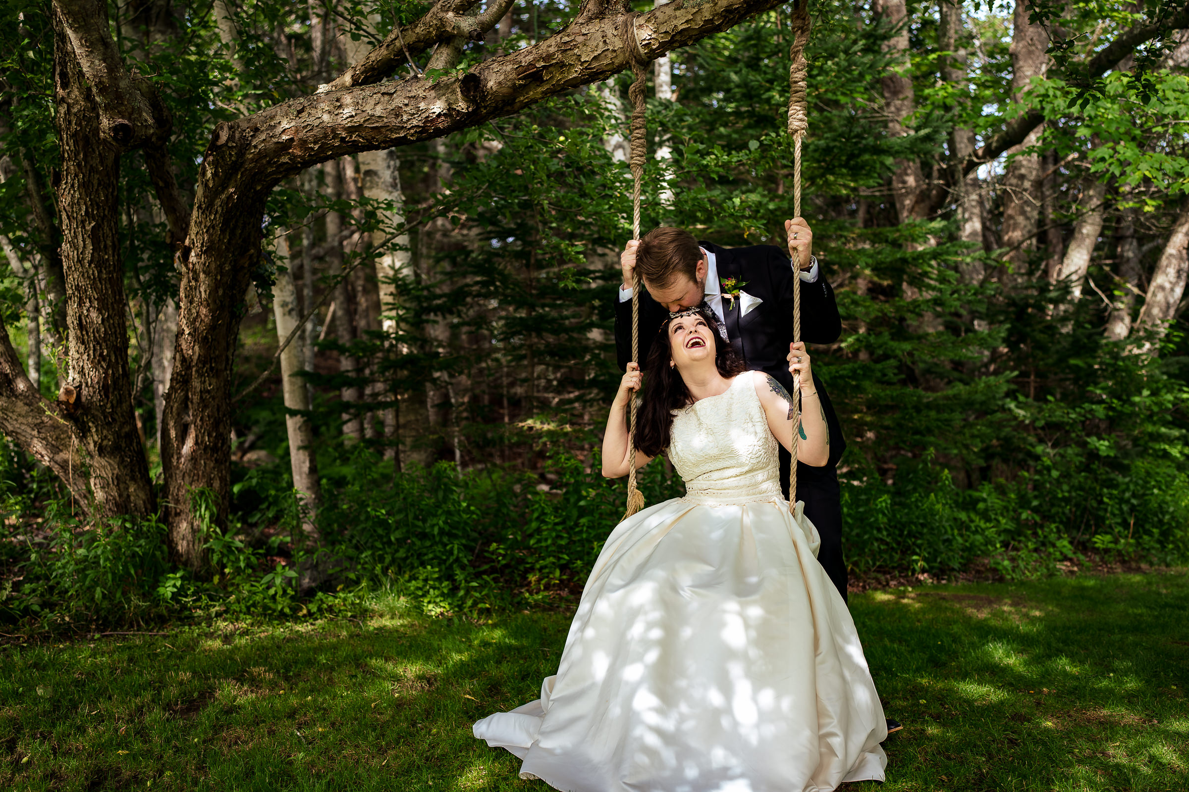 Bride sits on a wooden swing with the groom behind her kissing her on the forehead