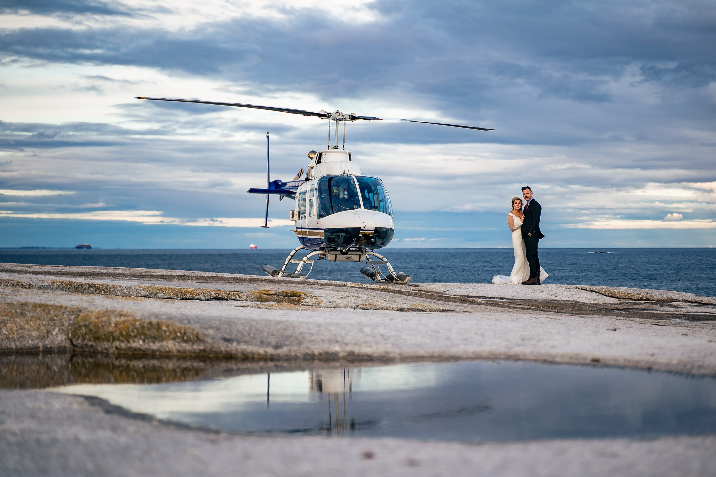 A bride and groom stand cuddling together in the distance next to a helicopter with the ocean behind them